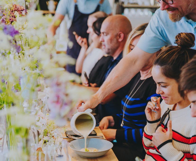 A person pours soup into a bowl on a table lined with people. The table is decorated with flowers and various bowls and utensils.