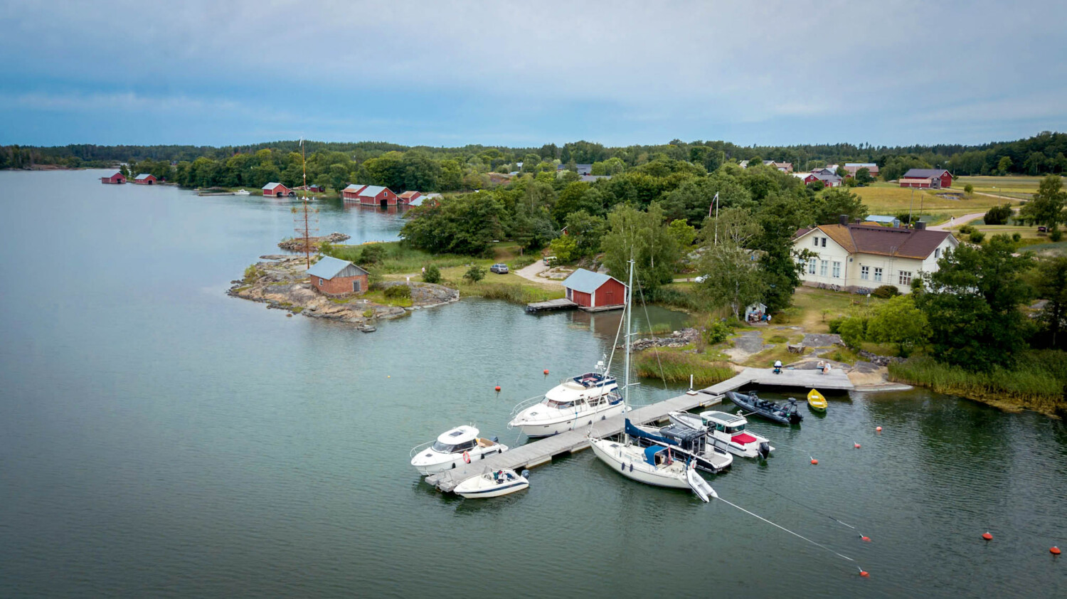 Aerial view of a small marina with several boats docked at a wooden pier, surrounded by a scenic landscape with trees, houses, and calm water.